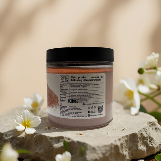 Himalayan salt scrub jar with moisturizing oils on stone surface, details on the back label visible, surrounded by flowers.