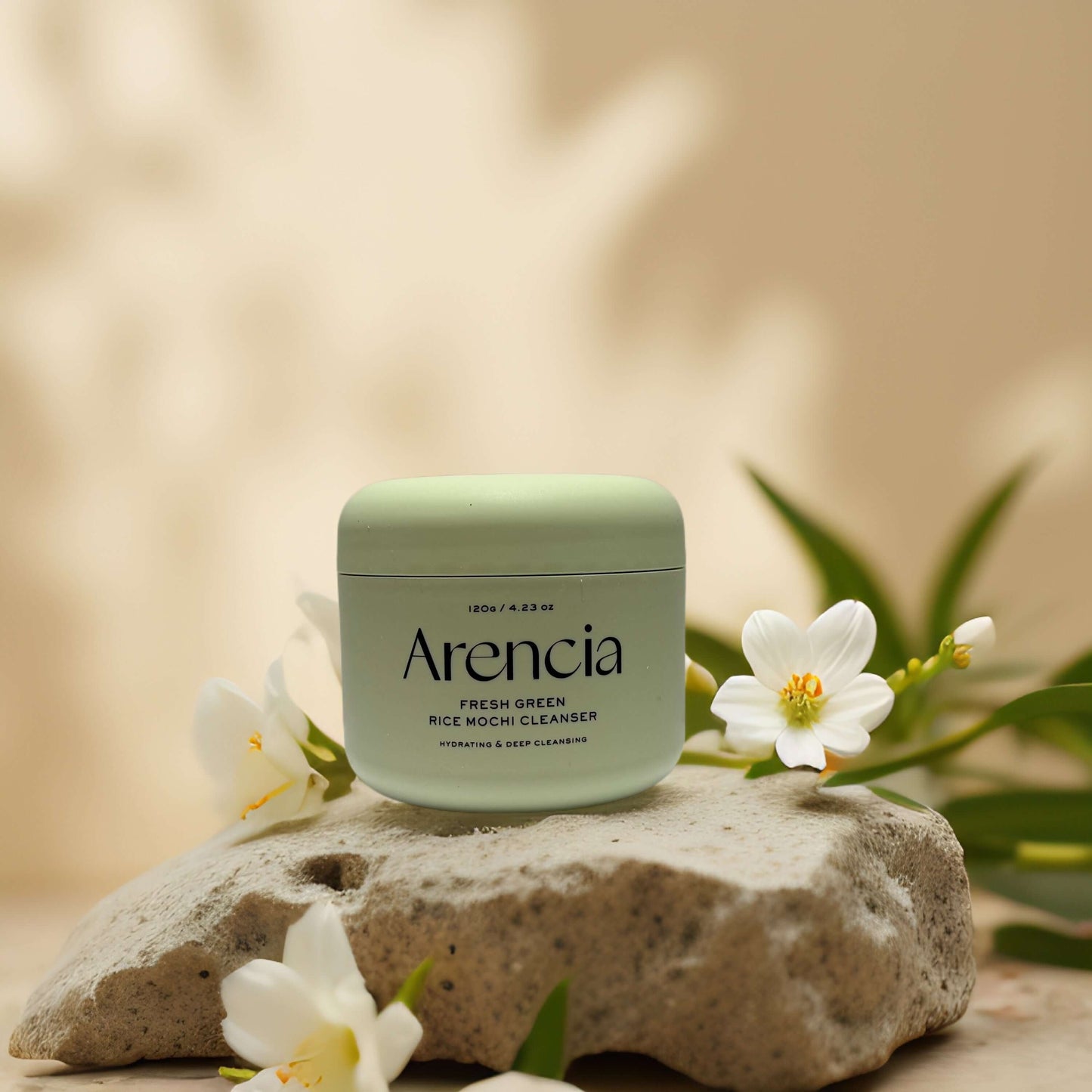 Arencia Fresh Green Rice Mochi Cleanser jar on a stone, surrounded by white flowers and greenery.