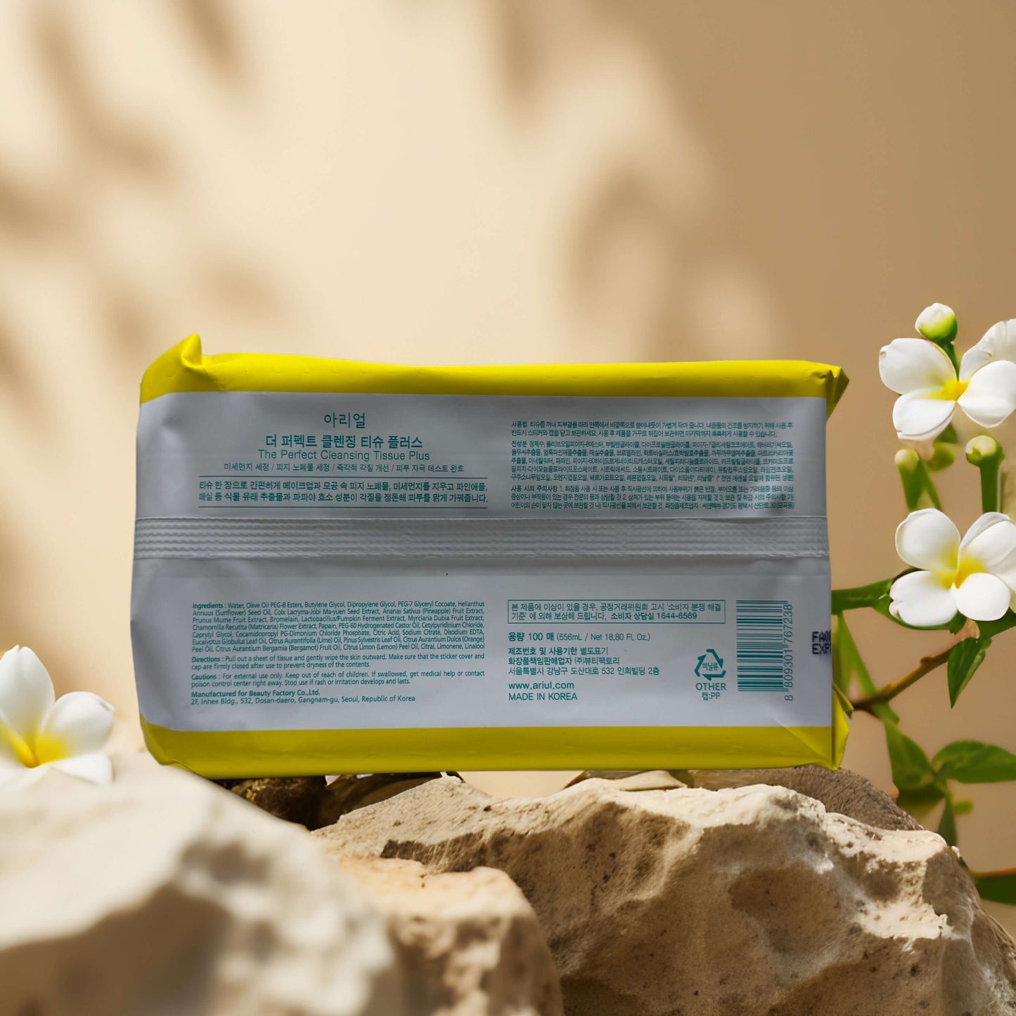 Ariul One-Step Facial Refresh cleansing tissue pack with yellow lid on rocks and surrounded by white flowers.