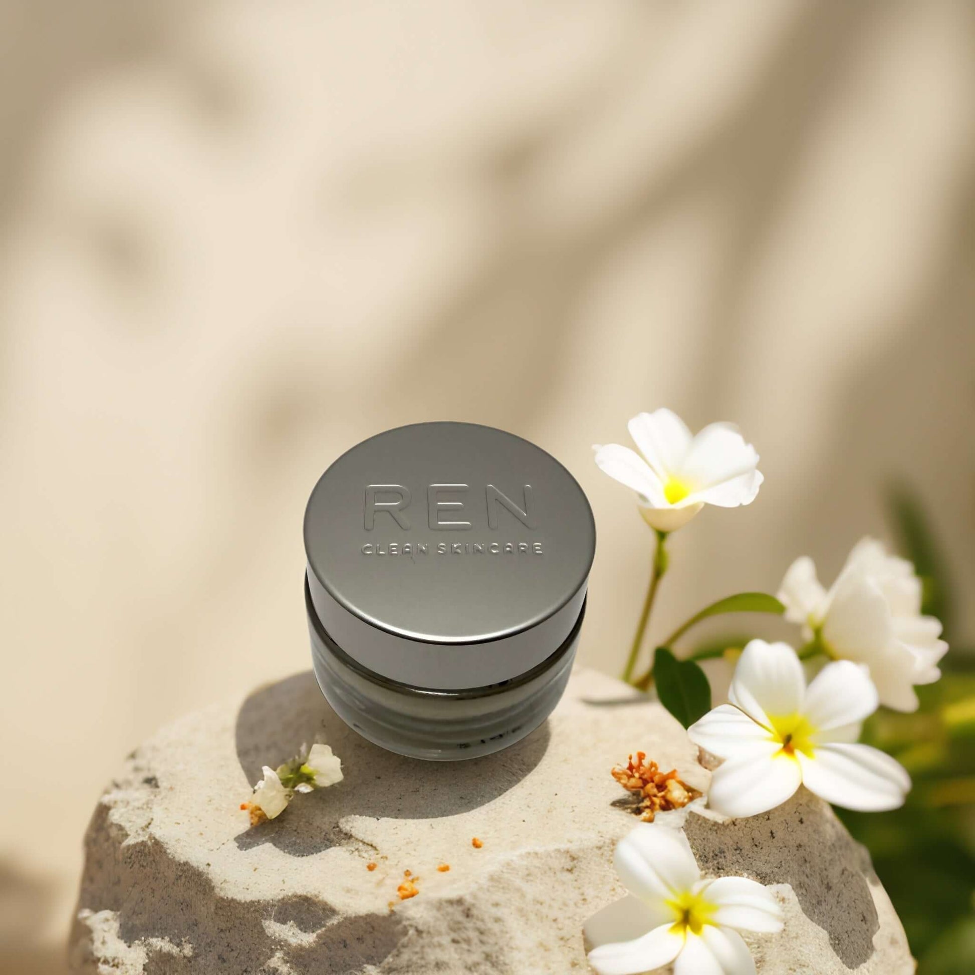 REN Global Protection Day Cream on a stone surface, surrounded by white flowers, ideal for sensitive skin care.
