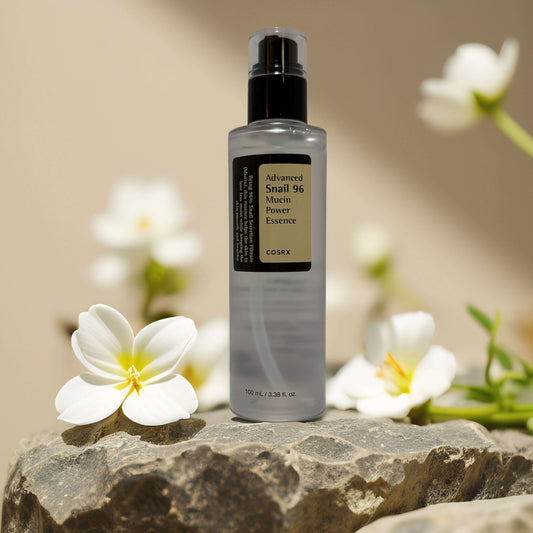 COSRX Advanced Snail 96 Mucin Power Essence bottle surrounded by white flowers on stone, promoting hydration and repair.