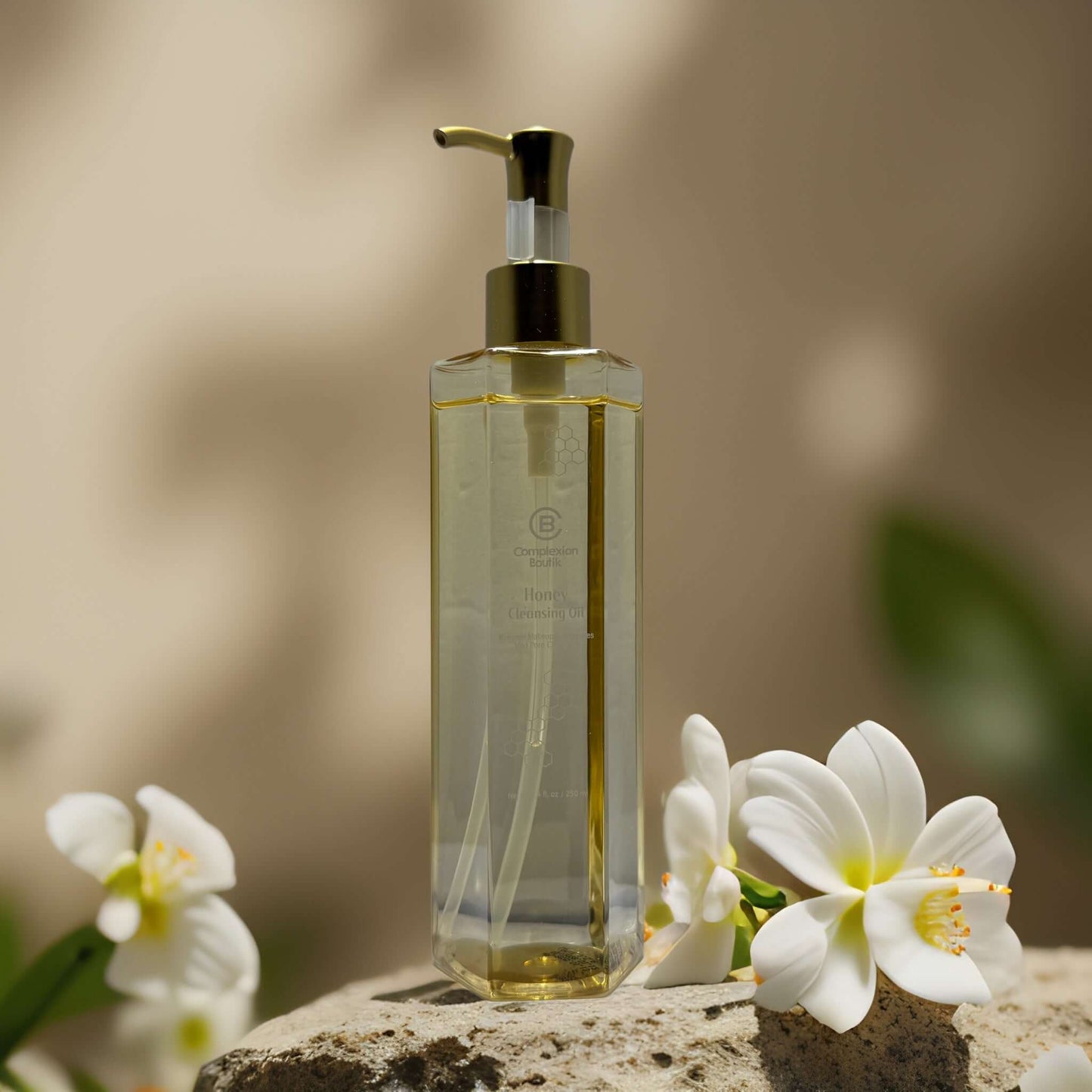 Honey Cleansing Oil by Complexion Boutik in a glass bottle with pump, surrounded by white flowers.