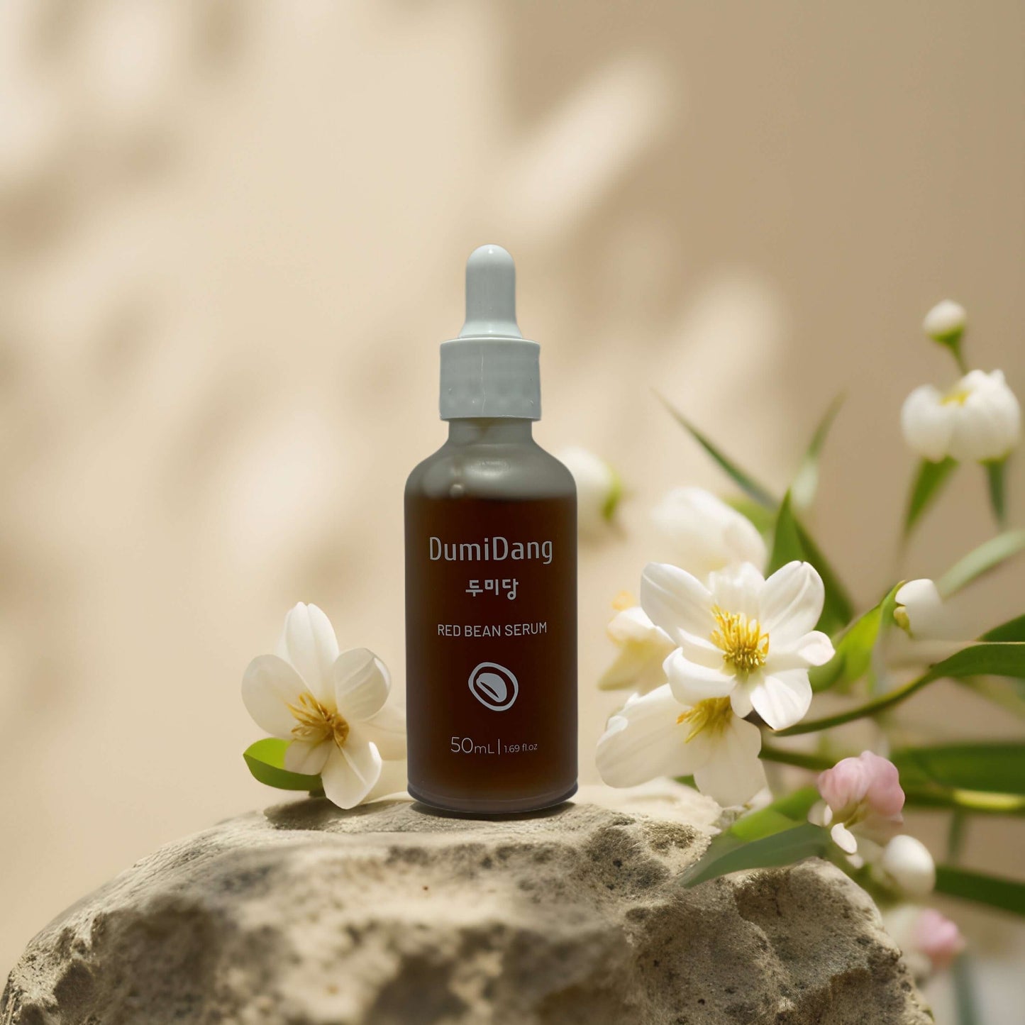 Dumidang Red Bean Facial Serum bottle on a stone, surrounded by flowers, promoting hydration and skin brightening.