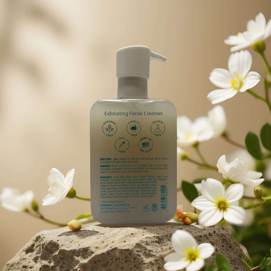 Gentle exfoliating facial cleanser bottle with pump, surrounded by white flowers on a rock