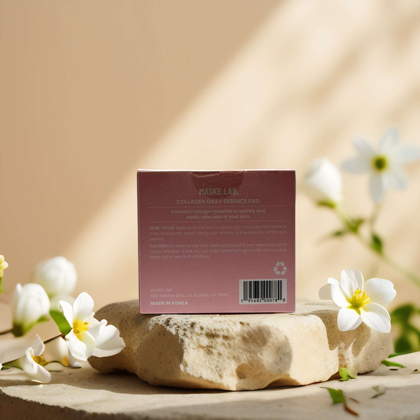 Collagen Essence Pads by Maske Lab packaging on a stone, surrounded by flowers, highlighting skincare benefits.