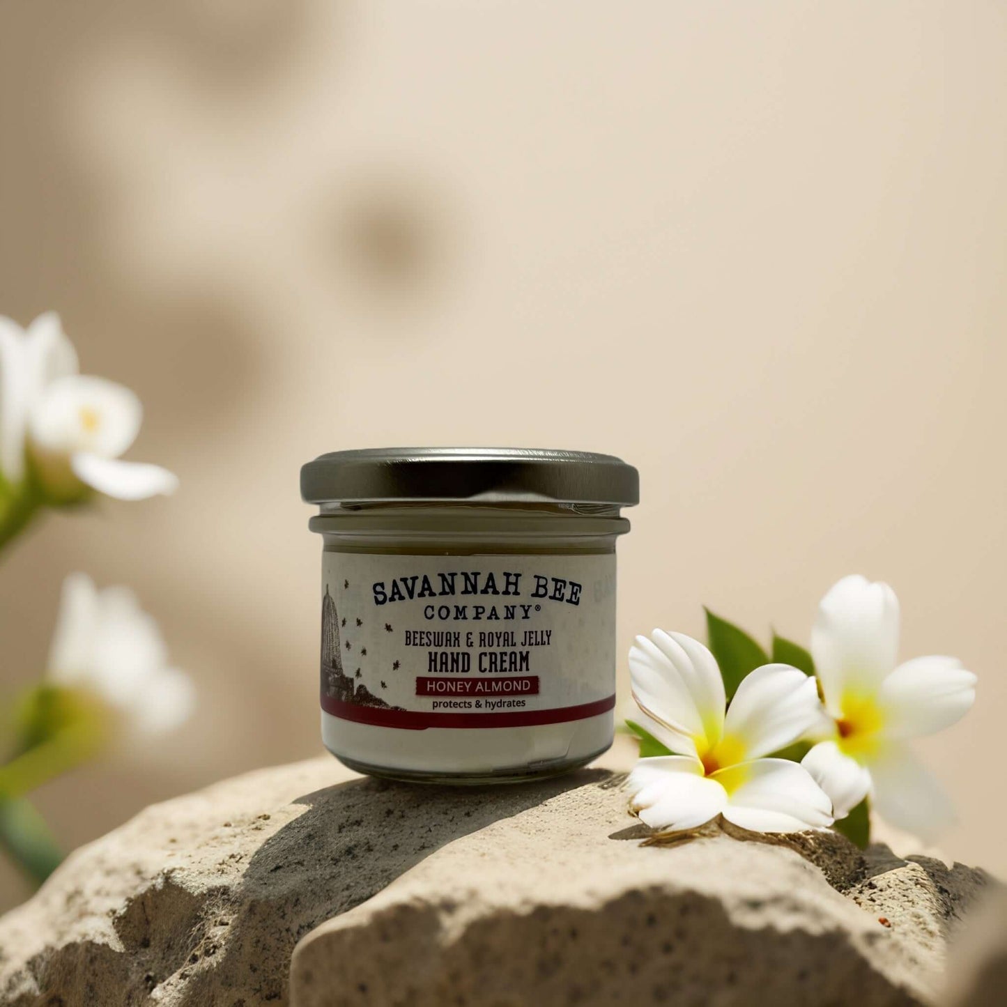 Savannah Bee honey almond hand cream jar on stone with white flowers, nourishing beeswax formula for dry hands.