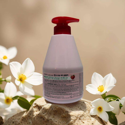 Welcos Vitalizing Hydration Strawberry Milk Body Cleanser bottle on a stone with white flowers, 19.8 fl oz.