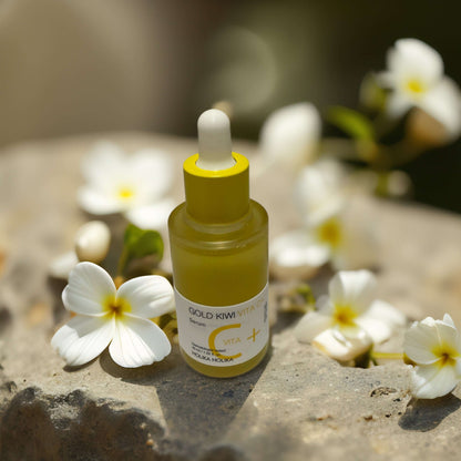 Gold Kiwi Serum by Holika Holika on stone with white flowers, promoting radiance and blemish reduction.