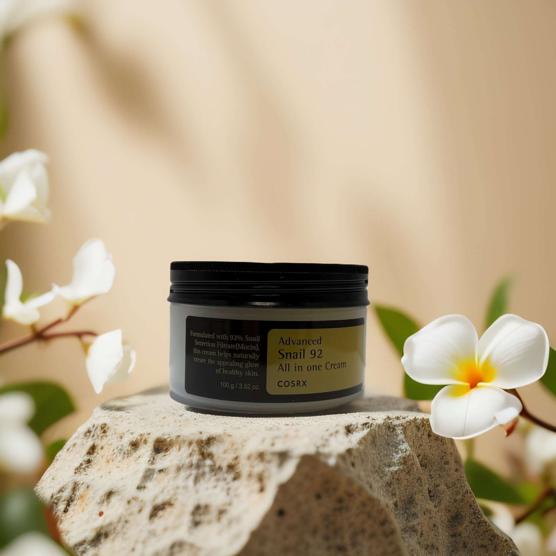 COSRX Advanced Snail 92 Face Cream on stone with flowers, 92% snail mucin for deep hydration and skin repair.