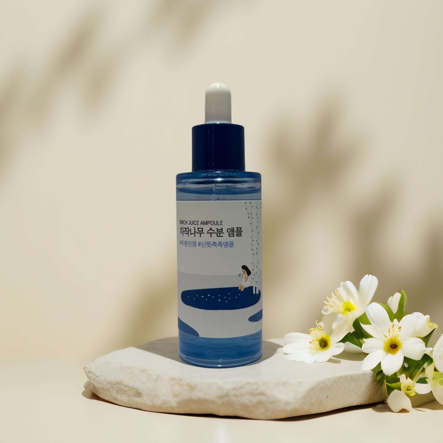 Round Lab Birch Juice Ampoule bottle on a rock with flowers.