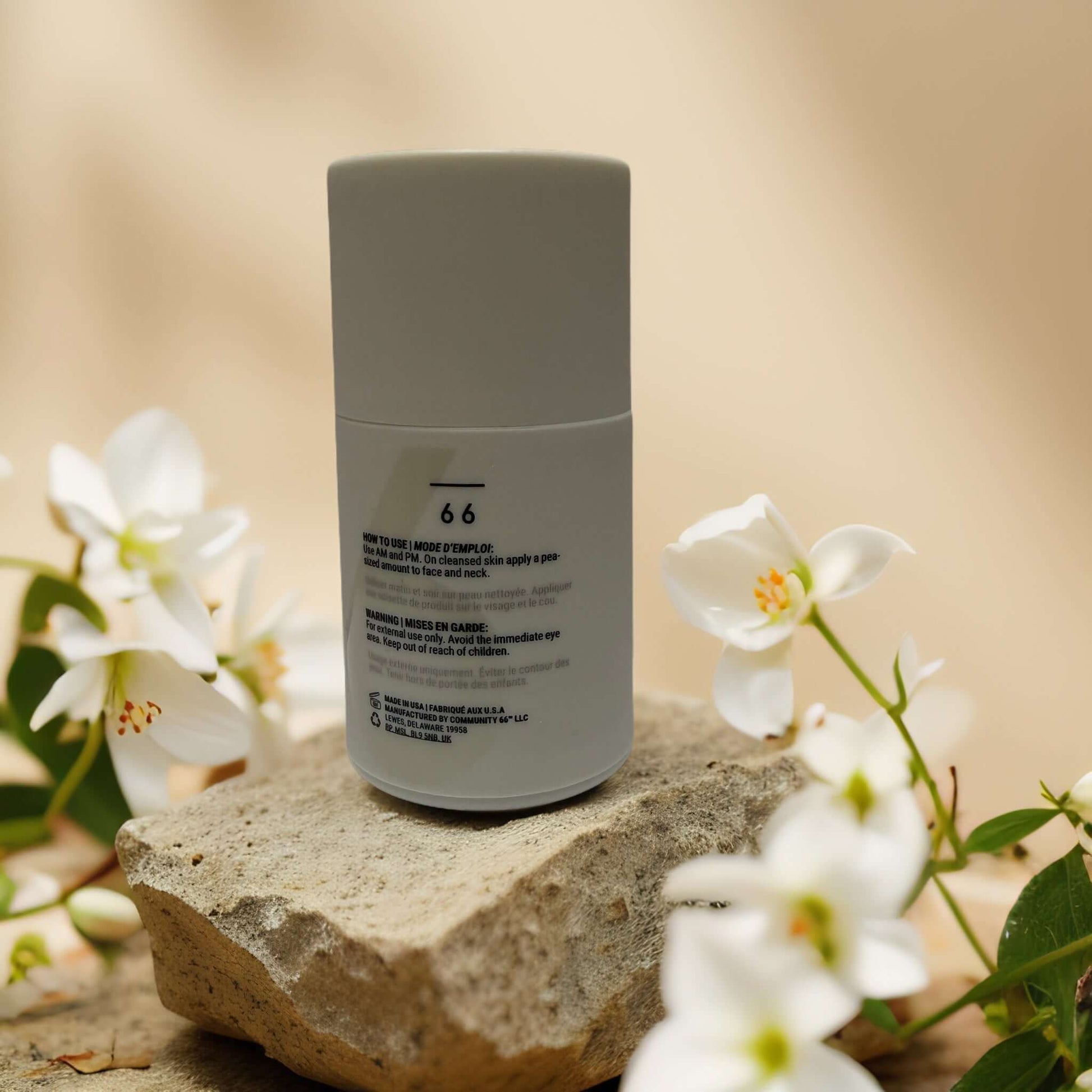 Back view of Community Sixty-Six Shine-Free Balance Oil Control Moisturizer bottle on a stone, surrounded by white flowers.